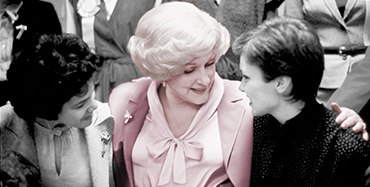 Mary Kay Ash stands between two women with her arms around them and listens as they are talking.